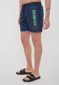 Maritime badkleding met een getailleerde tailleband, voorzien van grote witte en groene "Bikkembergs"-tekst aan de zijkant. Draagt met zwarte slipper sandalen.