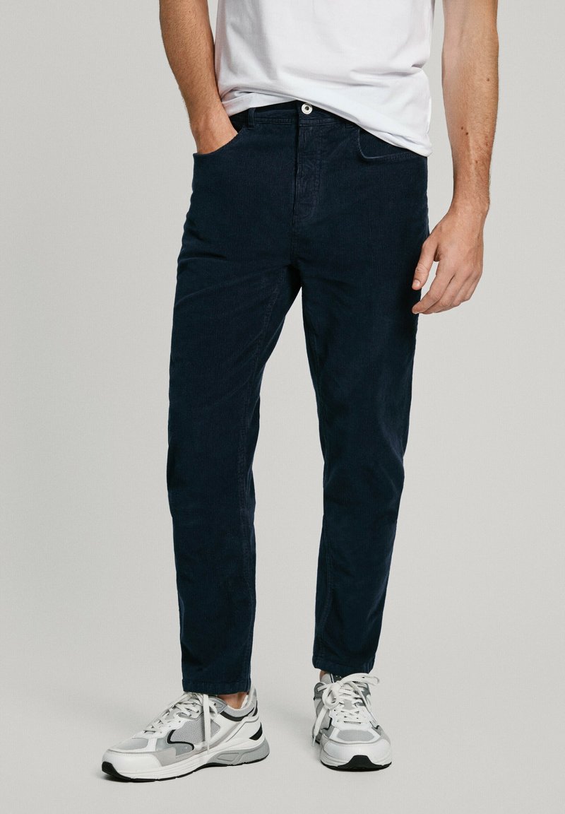 Homme portant un pantalon bleu marine foncé et des baskets blanches, debout avec une main dans la poche et un t-shirt blanc partiellement rentré.