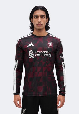 Hombre con camiseta de fútbol de Liverpool a cuadros granate y negro con rayas blancas de Adidas y logos de patrocinadores, de pie mirando al frente.