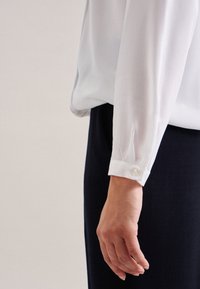 Blusa bianca con maniche lunghe, tessuto trasparente, polsino con dettaglio a bottone, abbinata a un pantalone di colore scuro. Texture liscia, design classico.