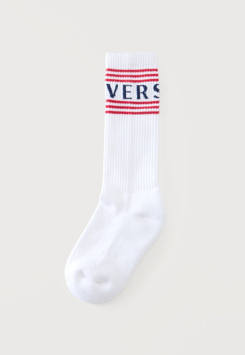 Chaussette blanche mi-mollet avec texture côtelée, présentant deux bandes rouges et des lettres bleues partiellement visibles près du haut.