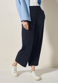 Zwarte wijde broek met een elastische tailleband, zijzakken en zachte stof, gecombineerd met witte sneakers en een lichtblauw shirt.