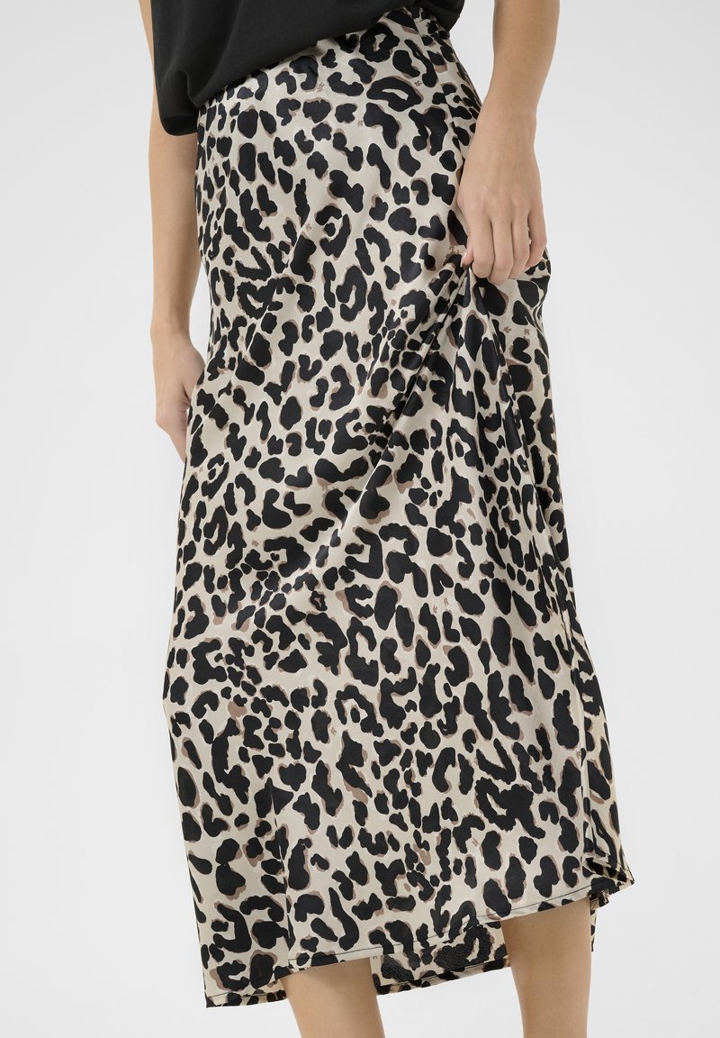 Femme portant une jupe midi imprimé léopard beige et noir, tenant un tissu sur un côté, avec un haut noir visible à la taille.