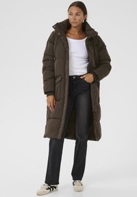 Lang, olivengrøn pufferjakke med hætte, der har lynlås, frontlommer og en quiltet design. Parret med mørke jeans og sneakers.