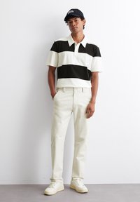 Kurzärmliges Polohemd mit schwarz-weißen horizontalen Streifen, kombiniert mit Cremefarbenen Hosen und weißen Sneakers; das Modell steht mit den Händen in den Taschen.