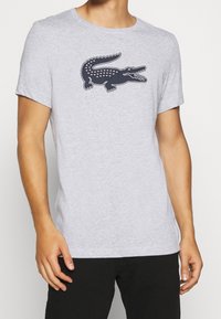 Grå kortärmad t-shirt i lätt tyg, med en mörkblå krokodil-logotyp på bröstet. Klassisk rund halsringning.