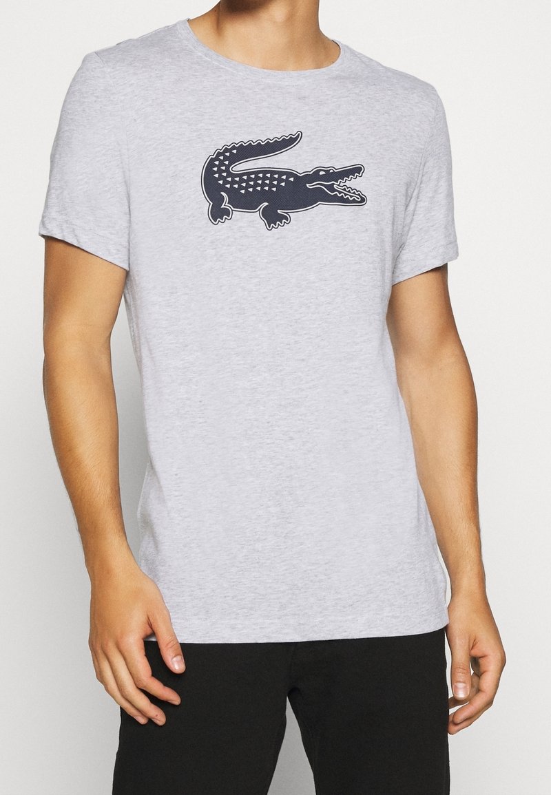 Grå kortärmad t-shirt i lätt tyg, med en mörkblå krokodil-logotyp på bröstet. Klassisk rund halsringning.