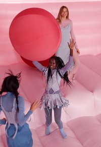 Trois personnes vêtues de tenues pastel s'amusent sur une surface gonflable rose. Une grande sphère rouge est lancée entre elles.