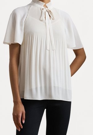 Femme portant un chemisier blanc à manches courtes avec des plis et un détail de nœud au col, associé à un pantalon noir.
