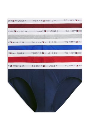 Ung mand, stående barfodet iført bordeauxfarvede briefs med hvid taljebånd på hvid baggrund, med en arm bag ryggen, vendt fremad.