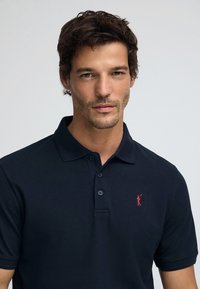 Mörkblå pikétröja med strukturerad yta, treknapparslå och liten röd broderad logotyp på vänster bröst. Normal passform.