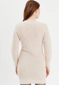 Vestido de punto en suave color beige. Presenta textura acanalada, mangas largas y cuello redondo. Largo hasta la rodilla con una silueta ajustada.