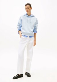 Felpa azzurra con logo frontale, indossata sopra una camicia a righe blu e bianche. I pantaloni bianchi a gamba larga e mocassini neri completano l'outfit.