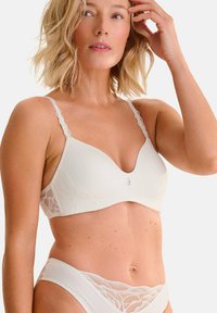 Soutien-gorge beige clair avec des bonnets souples, des panneaux latéraux en dentelle et des bretelles décoratives. Décolleté en V remarqué et détail logo subtil au centre.