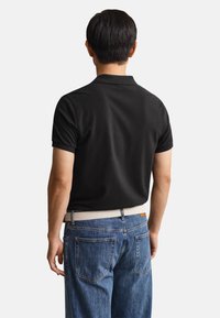 Man wearing black short-sleeve polo shirt tucked into blue jeans with a white belt, facing away on plain light background.