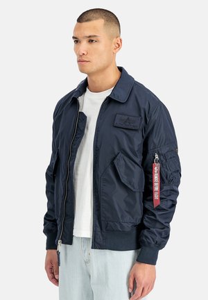 Male model wearing navy blue bomber jacket with front pockets, zip closure, and red "Remove Before Flight" tag on left sleeve pocket.