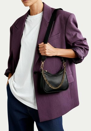 Osoba ubrana w fioletową marynarkę, białą koszulkę i ciemne jeansy, trzymająca czarną torebkę z złotym łańcuszkiem i paskiem na ramię.