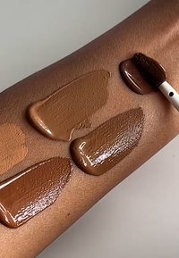 Fire prøver af flydende foundation i forskellige nuancer på en hudoverflade, der viser forskellige toner, samt en pensel, der påfører produktet.