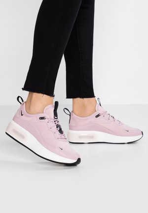 Persona vistiendo zapatillas deportivas de color rosa claro con suelas blancas gruesas y acentos negros, combinadas con pantalones negros recortados sobre un fondo sencillo.