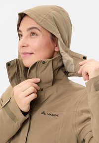 Persona che regola il cappuccio di una giacca beige della marca Vaude, dotata di zip e chiusure a scatto, guardando verso l'alto.