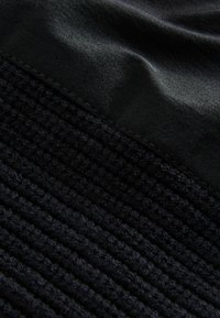 Tessuto nero composto da una trama intrecciata, con una texture a coste e una superficie liscia e lucida. Dettagli delle cuciture visibili nei punti in cui le texture si incontrano.