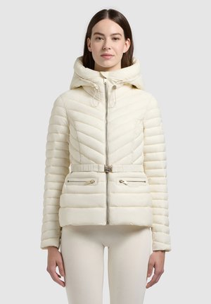 Kobieta w kremowej pikowanej kurtce z paskiem, suwakiem z przodu, kapturem i kieszeniami na zamki, stojąca na jednolitym jasnym tle.