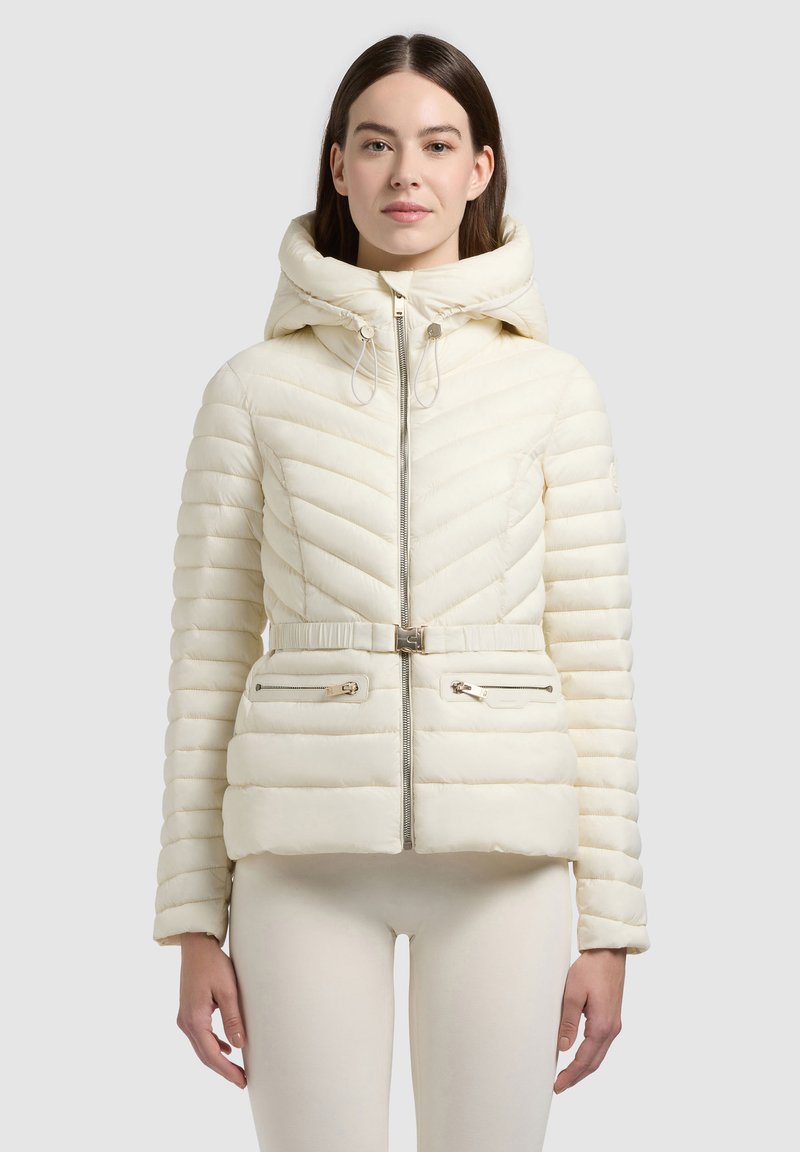 Femme portant une veste matelassée crème avec ceinture, fermeture éclair devant, capuche et poches zippées, debout devant un fond clair uni.