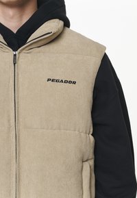 Béžová manšestrová vesta se zipem vpředu, s horizontálním prošitím a černým logem "PEGADOR". Nošena přes černou mikinu s kapucí.