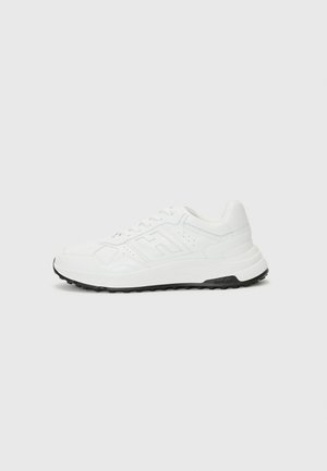 Chaussure de sport blanche avec un dessus synthétique texturé, dotée d'une semelle en caoutchouc noire contrastante et de détails perforés sur les côtés.