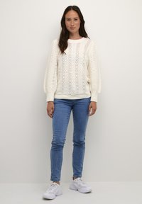 Pull en tricot blanc avec un motif de vagues, coupe décontractée et poignets côtelés ; associé à un jean skinny bleu clair et des baskets blanches.