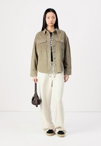 Olive green corduroy shirt jacket with two chest pockets and silver buttons, paired with cream wide-leg pants and black slip-on shoes.