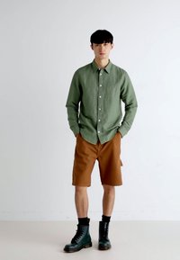 Camicia verde a pulsante, pantaloni corti marroni e stivali da combattimento neri. Tessuto leggero, vestibilità classica, pantaloni corti all'altezza del ginocchio, texture liscia.