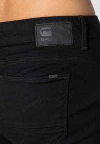 Jeans de mezclilla negra con una etiqueta de cuero que dice "RAW" en la cinturilla. Presenta un bolsillo trasero con una etiqueta más pequeña y detalles de costura sutiles.