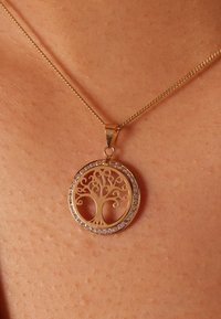 Collier en or avec un pendentif circulaire au design d'arbre entouré d'un anneau de pierres précieuses transparentes, présenté sur un ton de peau.