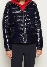 Osoba ubrana w błyszczącą granatową kurtkę puchową zapinaną na zamek, nosi czerwoną bluzę z kapturem i ciemnogranatowe spodnie, stojąca na jednolitym tle.