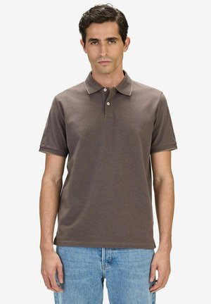 Man wearing a short-sleeve brown polo shirt and light blue jeans standing against a plain white background.