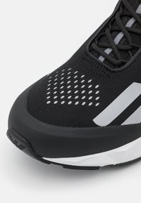 Svart sneaker-tå med vita prickiga ventilationsöppningar, svarta snörning, två reflekterande silverränder och vita geometriska detaljer på sulan.