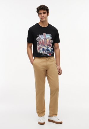 Ung mand iført sort Miami grafik T-shirt, beige bukser og hvide sneakers, stående med den ene hånd i lommen mod en hvid baggrund.