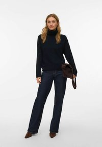 Jūras zils augsta kakla džemperis ar vaļēju piegriezumu un ribotiem manžetēm, kopā ar tumšām zvīņdiagona džinsa biksēm ar uzplaķēm un brūniem papēžu mulei, turot brūnu somu.