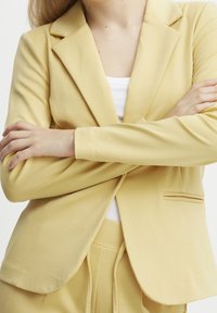 Blazer jaune à coupe ajustée, tissu doux, revers structurés et une poche poitrine unique; associé à un haut blanc.