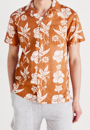 Homme portant une chemise orange à manches courtes boutonnée avec un imprimé floral blanc et un short gris clair à rayures verticales.