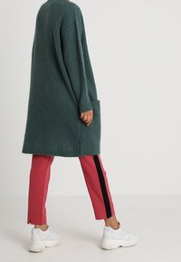 Cardigan lungo verde a coste con tasca, abbinato a pantaloni rosa con una striscia nera laterale e scarpe da ginnastica bianche.