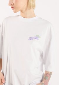 Λευκό βαμβακερό t-shirt με μια μικρή, πολύχρωμη γραφική εικόνα στην αριστερή πλευρά του στήθους, στρογγυλή λαιμόκοψη και κοντά μανίκια με άνετη εφαρμογή.