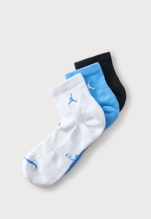 Trois chaussettes basses pliées en blanc, bleu et noir, chacune avec un petit logo près du bord-côte.