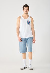 Maglietta senza maniche bianca in cotone con taschino a stampa tropicale, abbinata a pantaloni corti di jeans blu. Il modello indossa calze bianche e sneakers a strisce nere.