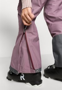 Vodotesné ružové lyžiarske nohavice so zipsovým bočným otvorom, sivými akcentmi na leme a čiernou lyžiarskou topánkou s viditeľným bielym nápisom.