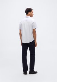 Άσπρο πουκάμισο με κοντά μανίκια και κυρτή διπλή ραφή στο κάτω μέρος, συνδυασμένο με σκούρο τζιν. Το πουκάμισο διαθέτει γιακά και διακριτικές λεπτομέρειες ραφής.