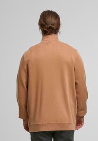 Hellbraunes Sweatshirt mit hohem Kragen, schlichtem Design, strukturiertem Stoff und gerippten Bündchen, getragen über dunkelgrauen Hosen.
