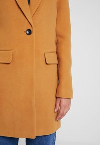 Abrigo de mezcla de lana color beige con un solo botón negro, mangas largas, dos bolsillos frontales y un diseño recto. Lleva puesto sobre unos vaqueros azules.