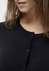 Cardigan in maglia nera con scollatura rotonda, dotato di due bottoni neri e texture a costine lungo i bordi. Inquadratura ravvicinata del tessuto.
